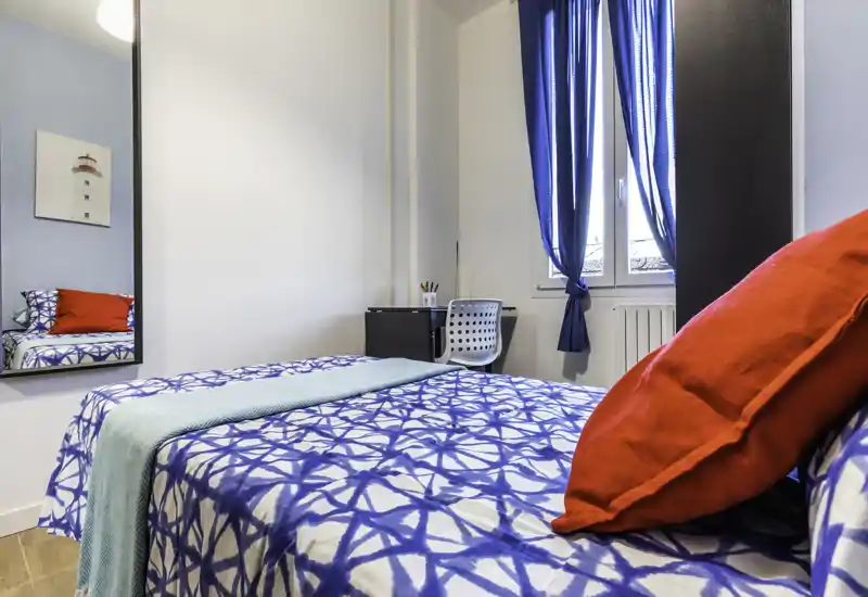 Camera da letto compatta e luminosa con copriletto blu a motivo, cuscino rosso e una piccola scrivania vicino alla finestra — ideale per un singolo che cerca un ambiente accogliente e ben illuminato.