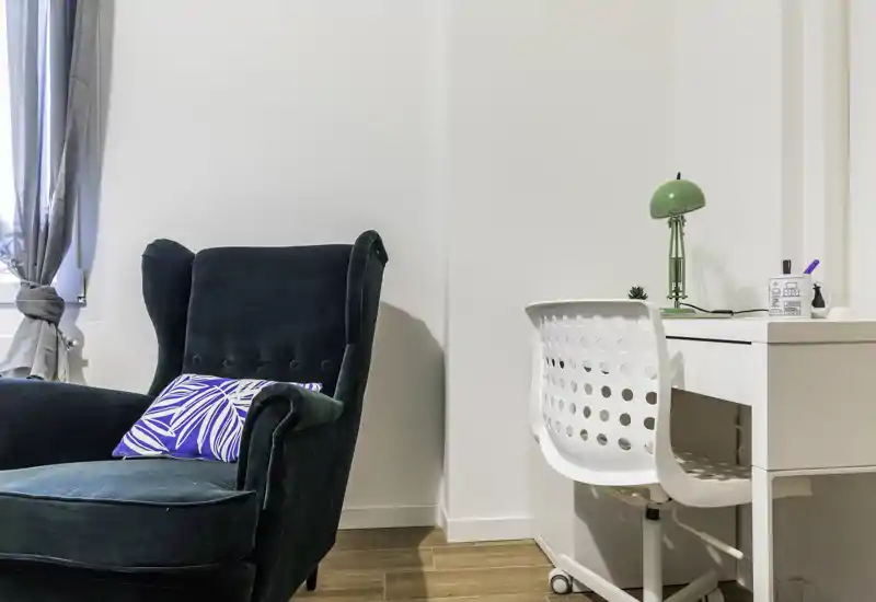 Angolo luminoso e di stile con una poltrona imbottita verde scuro e una scrivania bianca compatta: ideale per rilassarsi e lavorare leggermente. La luce naturale dalla finestra e gli accessori creano un ambiente accogliente e funzionale.