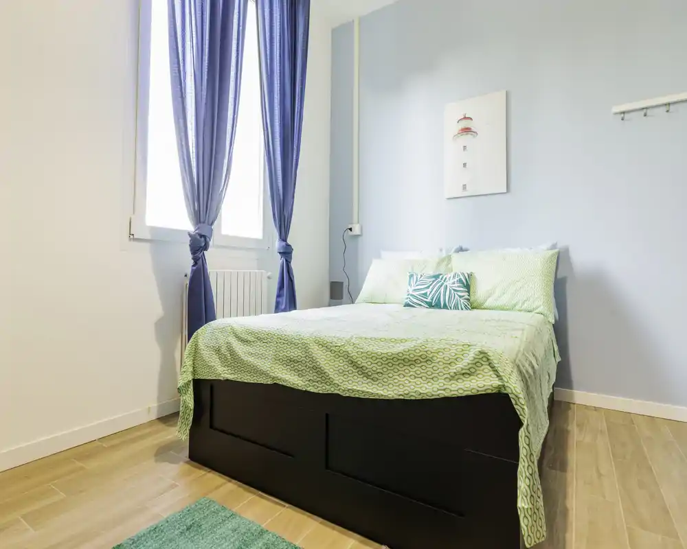 Bright, neatly styled bedroom with a double bed dressed in green bedding, blue curtains and light wood flooring, offering a fresh coastal feel.
