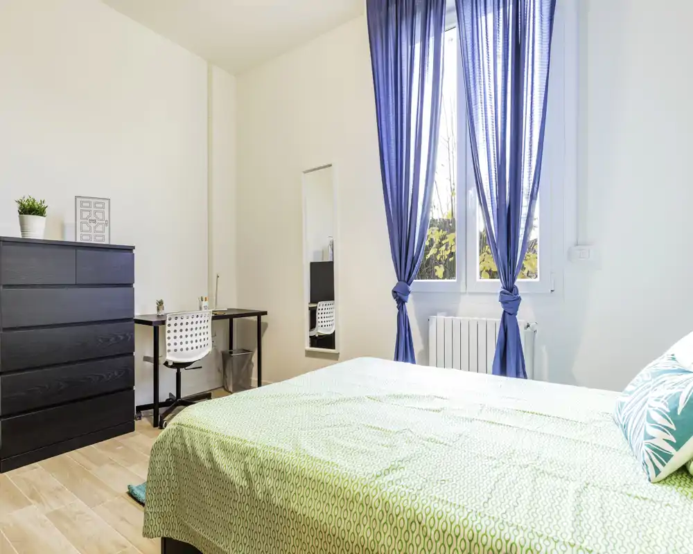 Bright, modern bedroom with a large bed in the foreground, blue window curtains and a small desk and chest of drawers — ideal for a comfortable stay.