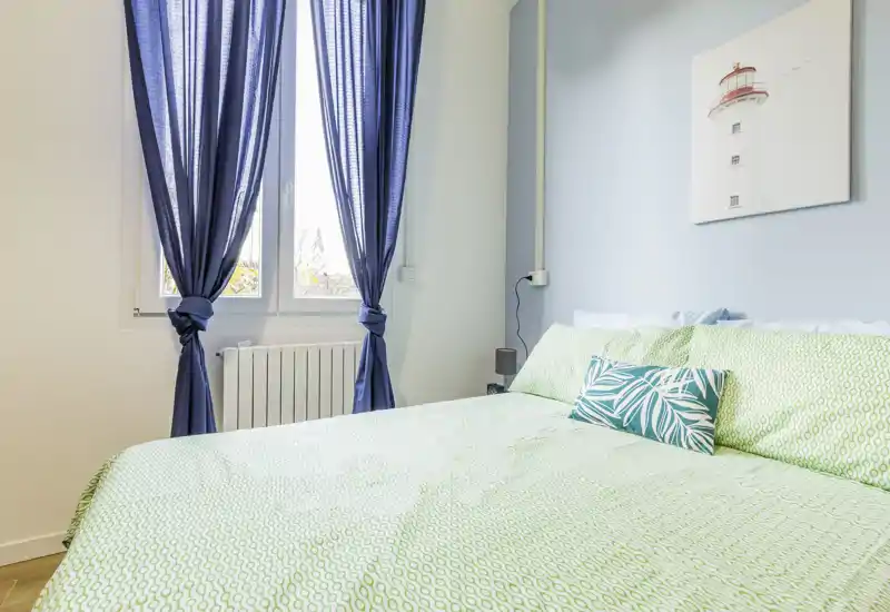 Bright, well-styled bedroom with a large bed dressed in green patterned linens, decorative cushions and natural light from the window with blue curtains.