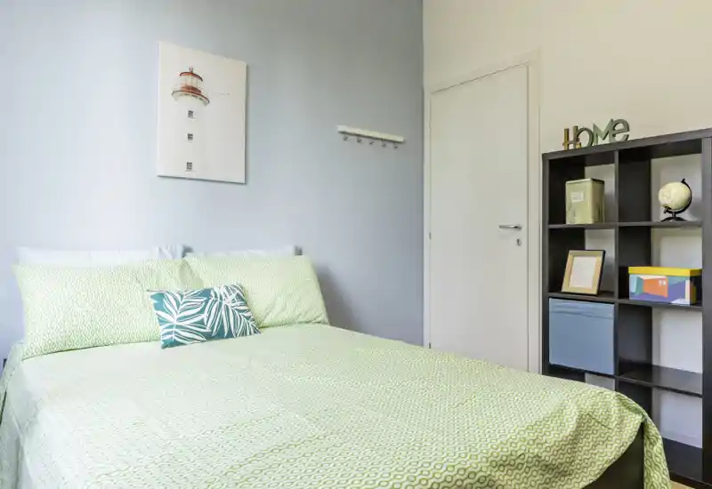 Bright, simply styled bedroom with a large bed dressed in patterned green linens, decorative cushions and a shelving unit for storage — ideal for a cozy short-stay.