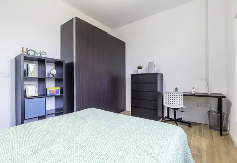 Bright, modern bedroom with a large bed in the foreground, dark wardrobe, shelving and a small desk — minimal, clean styling ideal for rentals.
