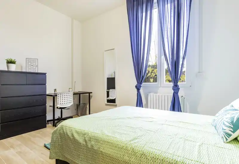 Bright, modern bedroom with a large bed in the foreground, blue window curtains and a small desk and chest of drawers — ideal for a comfortable stay.