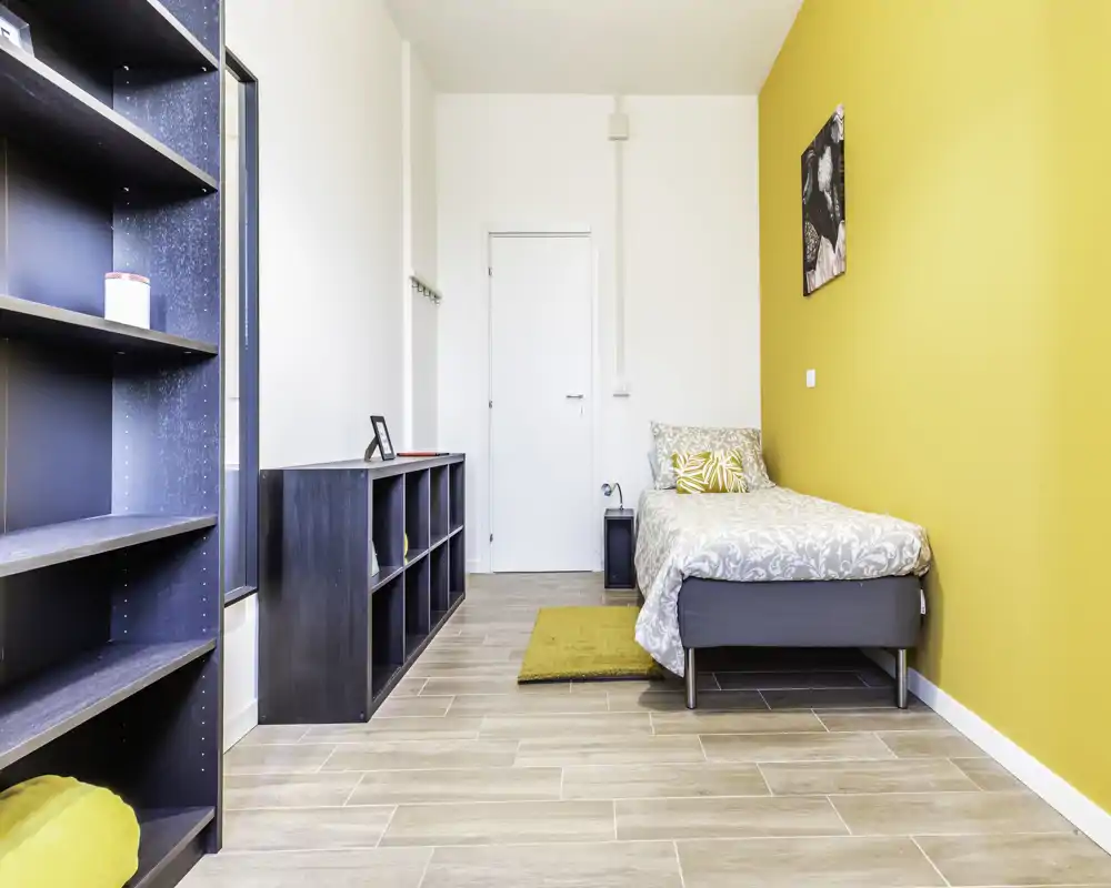 Chambre simple moderne et lumineuse avec lit confortable contre un mur jaune chaleureux, étagères bien rangées et sol clair effet bois — idéale pour une location cosy.