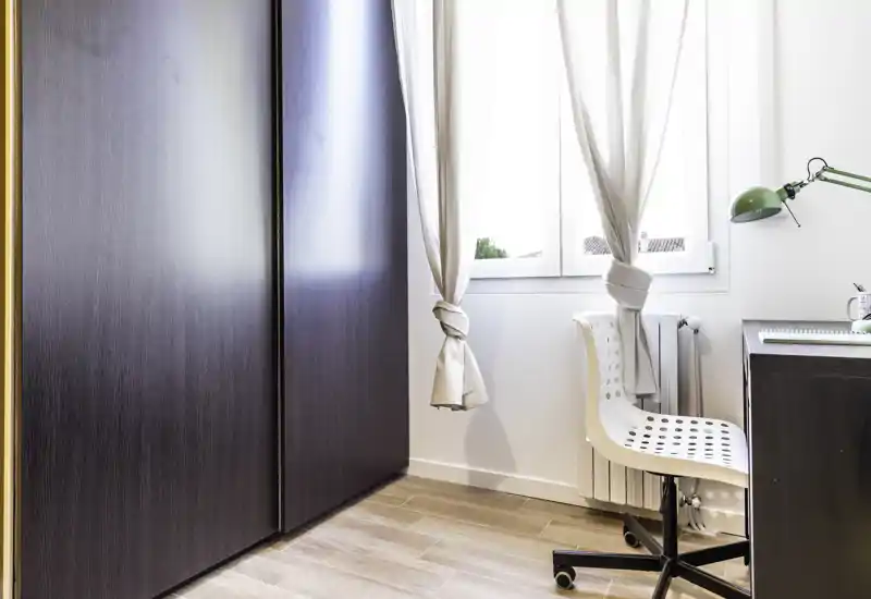 Angolo luminoso e minimale con armadio, finestra e una piccola scrivania/sedia da ufficio — un'area compatta soggiorno/studio con luce naturale.