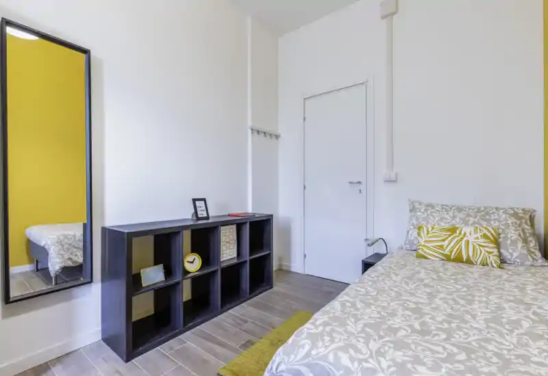 Camera da letto singola luminosa e curata con biancheria a motivo e cuscino giallo di accento; presente una libreria a cubi e uno specchio a figura intera che amplia lo spazio.