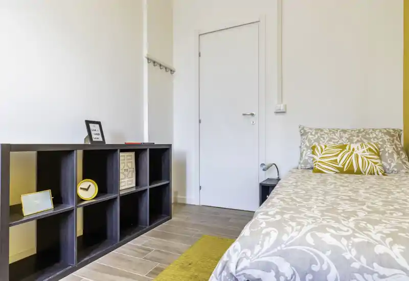 Camera singola luminosa e moderna con copriletto decorato, cuscino giallo e libreria a cubi bassa; linee pulite e pavimento effetto legno creano un ambiente accogliente e ben illuminato.