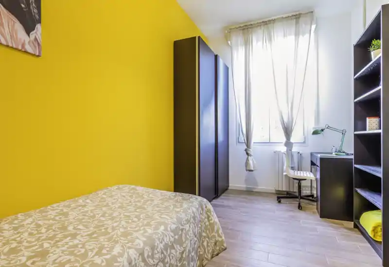 Camera da letto luminosa e ordinata con parete accentuata in giallo, letto singolo con copriletto decorato, armadio e una scrivania vicino alla finestra che lascia entrare molta luce naturale.