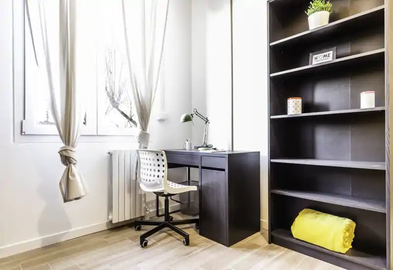 Angolo camera luminoso e compatto con scrivania e libreria — ampia finestra che illumina, lampada da tavolo moderna e sedia girevole per uno spazio lavoro funzionale.