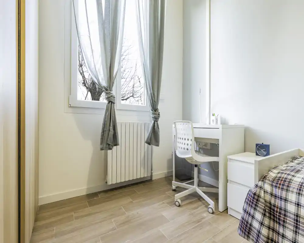 Angolo di camera da letto luminoso con letto singolo parzialmente visibile, scrivania bianca e sedia vicino a una grande finestra che permette molta luce naturale. Arredamento minimal e pavimento in legno chiaro.