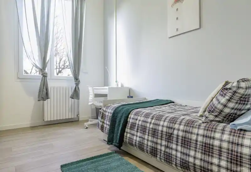 Bright, tidy single-bedroom with a plaid bed, green accents and a small desk by the window — ideal for a student or solo renter.