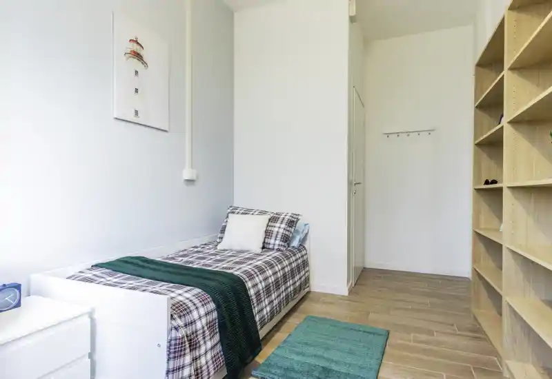 Bright, neatly styled single bedroom with a comfortable bed, plaid bedding, wooden shelving and tasteful accents — ideal for a cosy rental room.