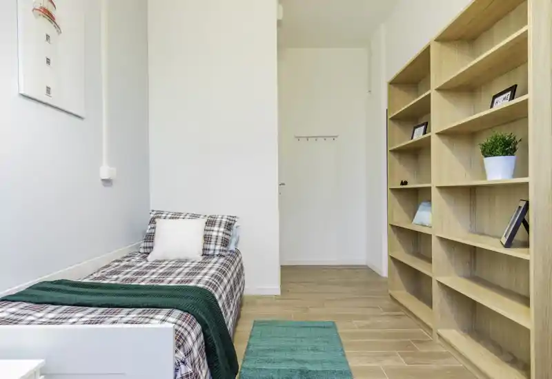 Bright, neatly styled single bedroom with a plaid-covered bed, green throw and rug, and built-in wooden shelving — a compact, cozy space ideal for one person.