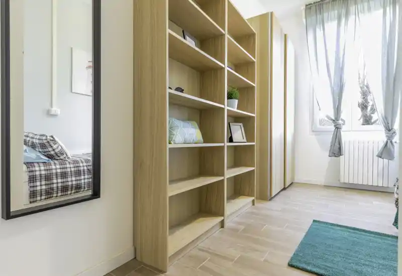 Bright, modern bedroom corner with light wood shelving, a mirror reflecting part of the bed, and a large window with sheer curtains providing soft natural light.