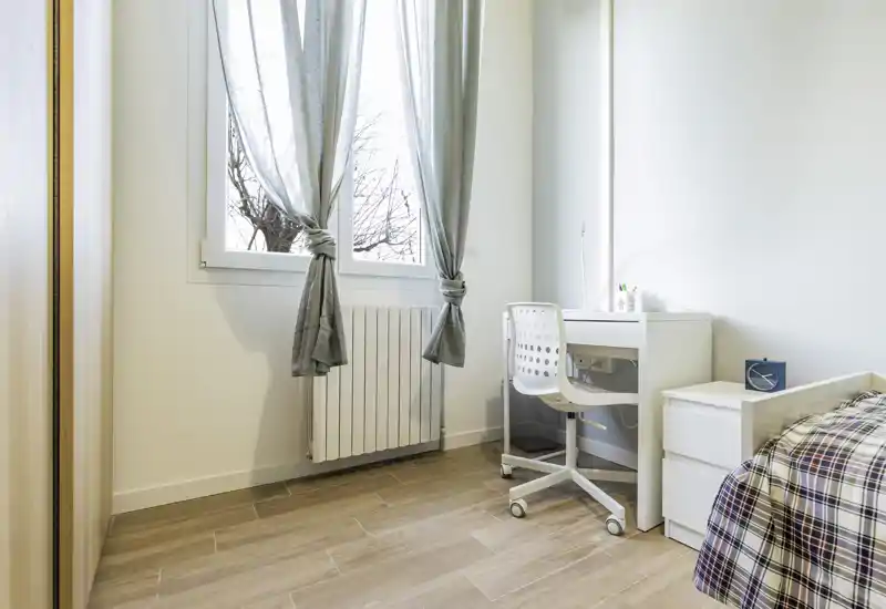 Small bright bedroom corner with a single bed partly visible, a white desk and chair by a large window letting in natural light. Minimal, modern decor with light wood flooring.