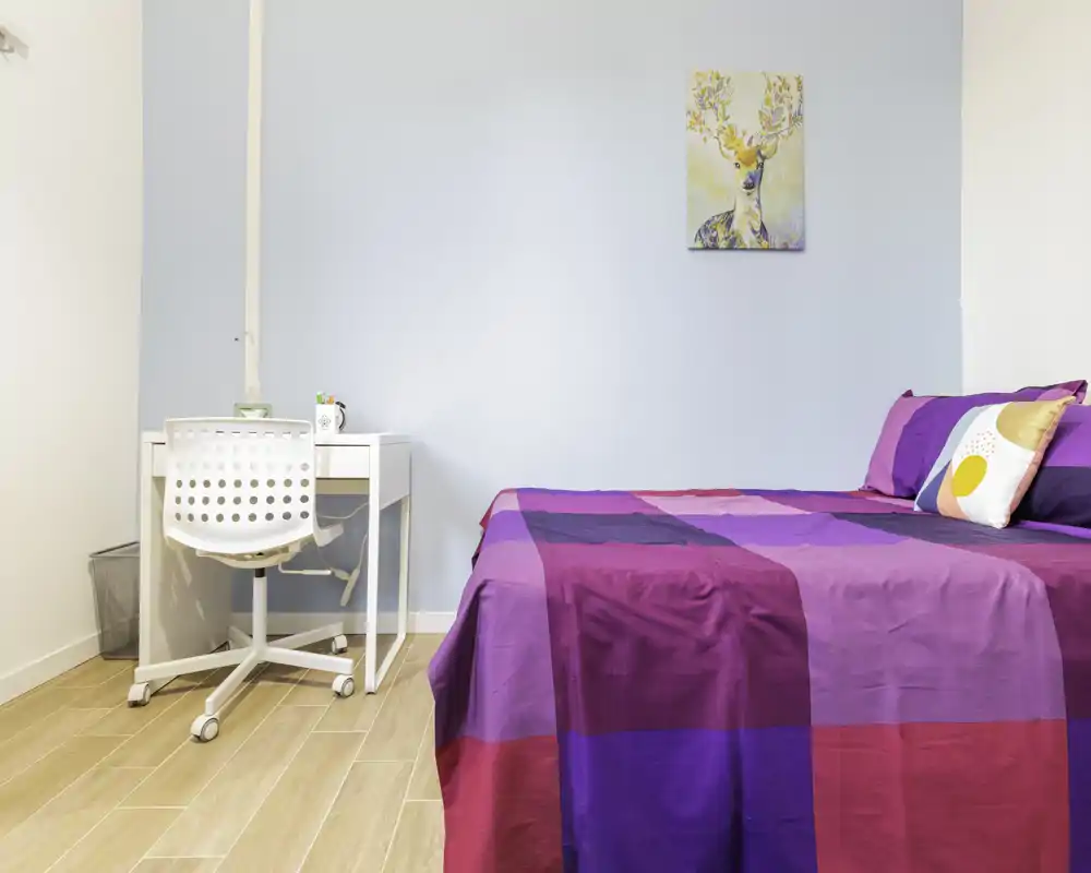 Camera da letto luminosa e curata con copriletto viola-rosa vivace, scrivania bianca e sedia: ideale per una persona o uno studente.