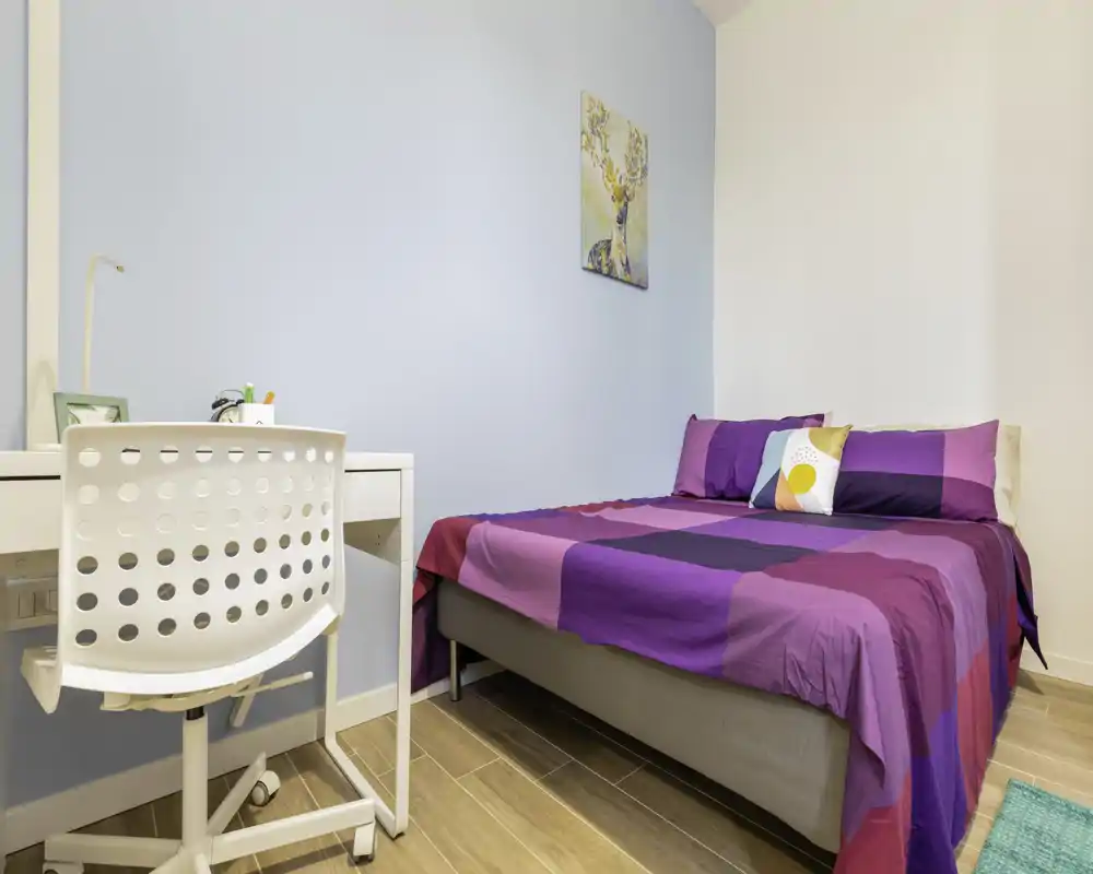 Camera da letto luminosa e ordinata con letto matrimoniale, biancheria viola colorata, una piccola scrivania e sedia e una parete accentata azzurra — ideale per una o due persone.