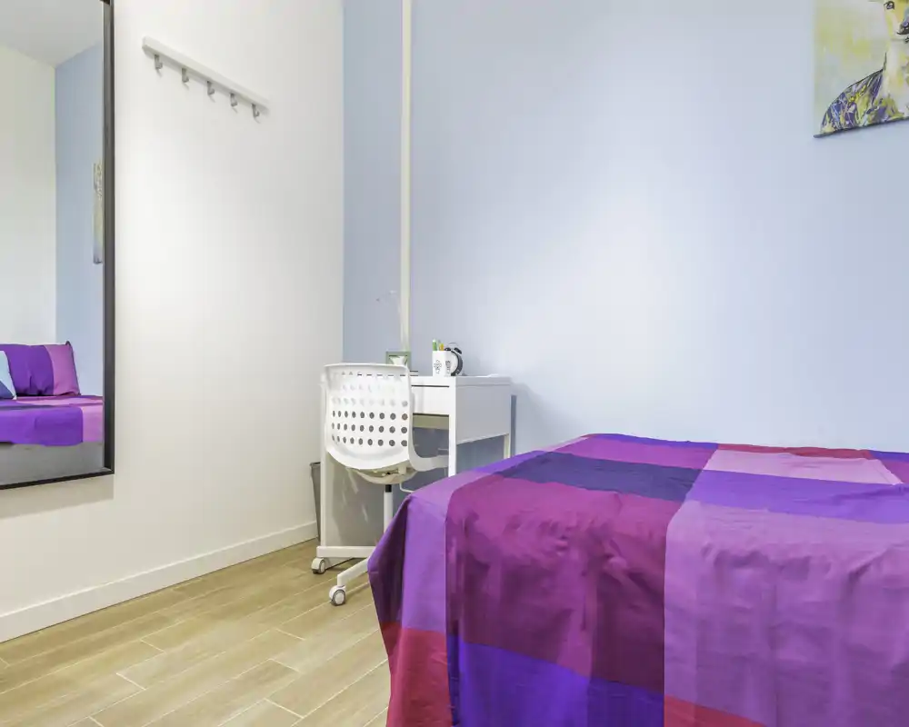 Camera singola luminosa e ordinata con una coperta colorata viola-fucsia, una piccola scrivania bianca con sedia e pavimento in legno chiaro. Arredamento essenziale e ben illuminato, perfetta per un inquilino singolo o un ospite.