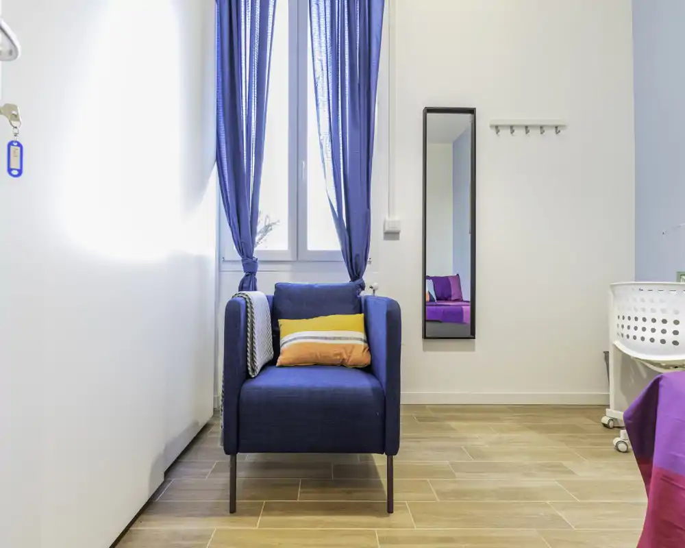 Graziosa area conversazione luminosa con poltrona blu sotto una finestra alta e cuscini colorati, perfetta per rilassarsi o leggere.