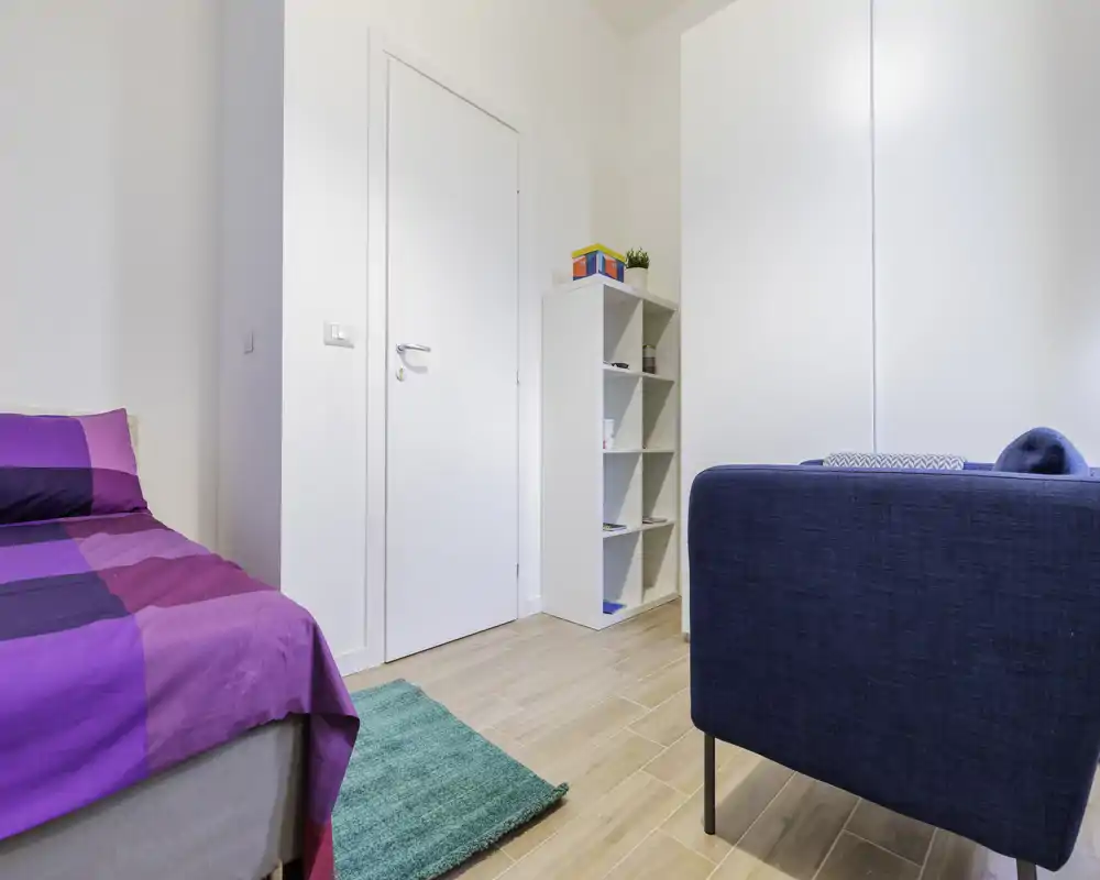 Piccolo angolo camera da letto luminoso con letto viola, poltrona imbottita blu, piccola libreria e tappeto verde su pavimento in legno — semplice, moderno e ben illuminato.