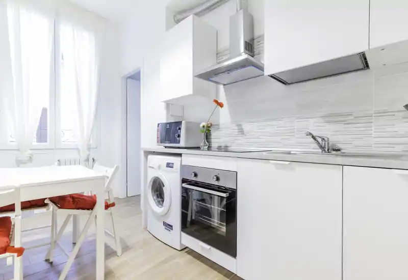 Bright modern kitchen with integrated appliances, a washing machine and oven, white cabinetry and a small dining area with red-cushioned chairs — ideal for a compact city apartment.