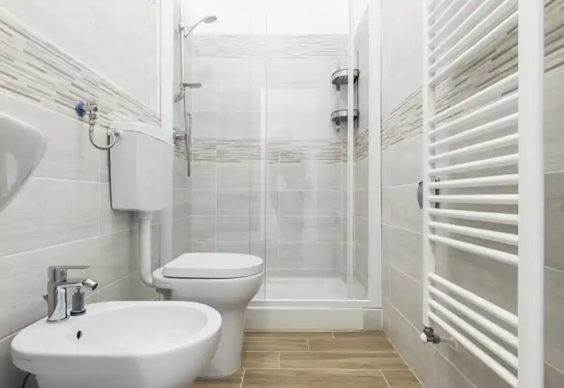 Bright, modern bathroom with shower cubicle, toilet, bidet and wall-mounted towel radiator. Neutral tiles and wood-effect floor create a clean, contemporary look.