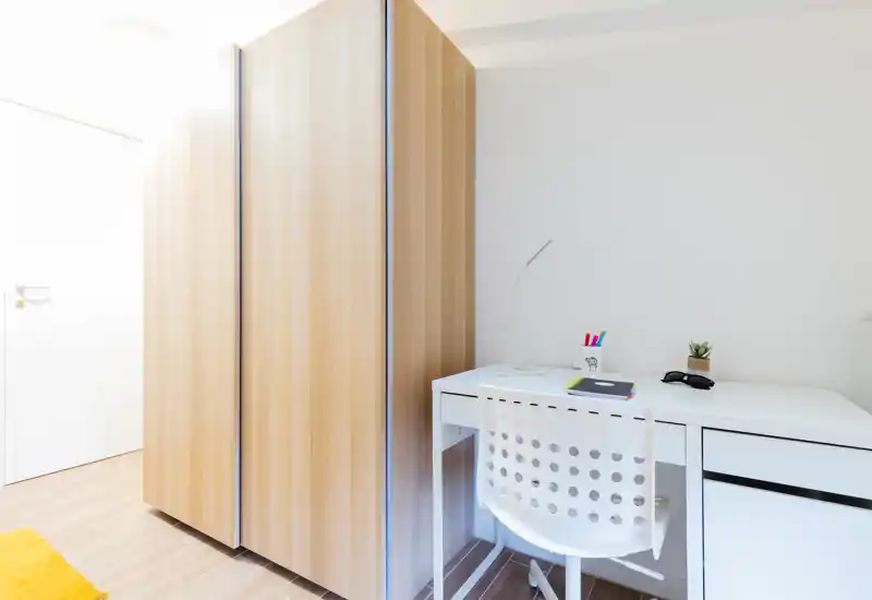 Bright, minimal bedroom corner with a large wooden wardrobe and a white desk with chair, lamp and small plant—clean, functional space ideal for a student or short-term stay.