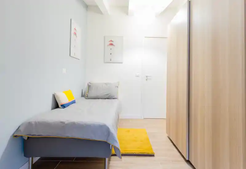 Sunny single bedroom with modern light-wood wardrobe, grey bedding and a cheerful yellow rug — a compact, well-lit room ideal for one person.