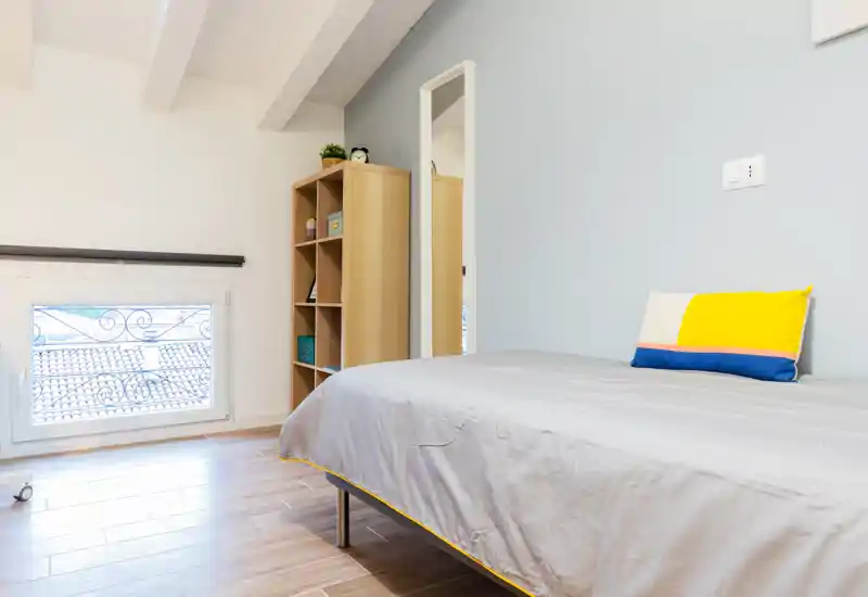 Bright, modern bedroom with a single bed, light wooden shelving and natural light from a low window — minimal, tidy and well lit for rental listings.