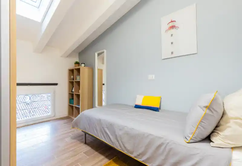 Bright, neatly styled single bedroom with a skylight and a comfortable bed dressed in grey linens and colorful cushions; light wood shelving adds storage and the pale blue wall with artwork creates a calm atmosphere.