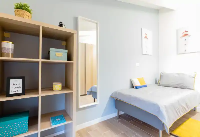 Bright, neatly styled single bedroom with a comfortable bed, decorative cushions, framed wall art and open shelving — ideal for a cozy rental.