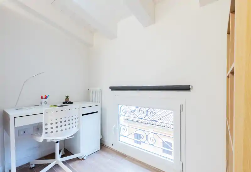 Bright, minimal home office corner with a compact white desk, perforated swivel chair, desk lamp and a small plant by a low window that fills the room with natural light.