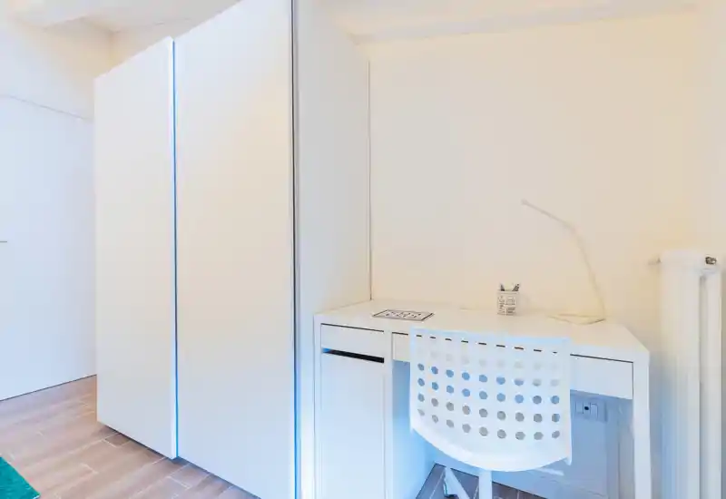 Small, bright bedroom corner featuring a white wardrobe and a compact desk with chair and lamp — minimal, modern furnishings ideal for a single occupant.
