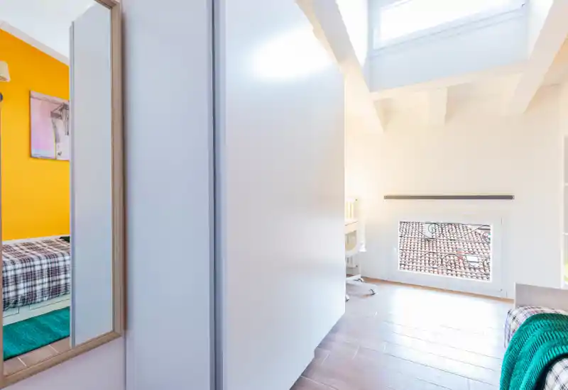 Bright, modern bedroom corner with a skylight and a small window overlooking tiled roofs. The shot includes a wardrobe and partial views of a bed with colourful textiles, ideal for a cozy single or guest room listing.