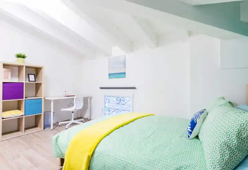 Dormitorio luminoso y bien decorado con una cama amplia vestida con ropa de cama verde y una manta amarilla; incluye escritorio y estantería, ideal para una estancia cómoda.