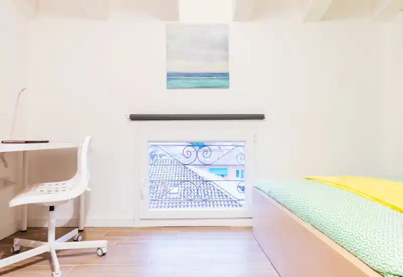Bright, minimal bedroom with a low wooden bed, colorful bedding and a small workspace by the window — ideal for a single or short-term stay.