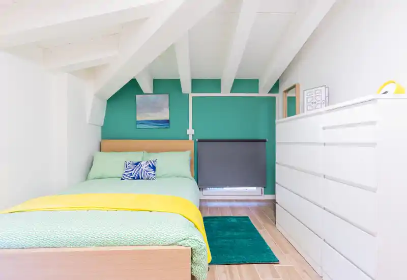 Dormitorio luminoso y bien presentado con cama completa, textiles coloridos y una cómoda blanca bajo techo abuhardillado con vigas.