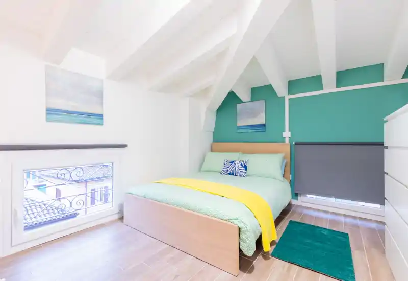 Dormitorio en ático, luminoso y con estilo, con cama doble de madera, ropa de cama colorida y pared de acento turquesa. El techo inclinado con vigas blancas y la luz natural generan una atmósfera fresca y acogedora.