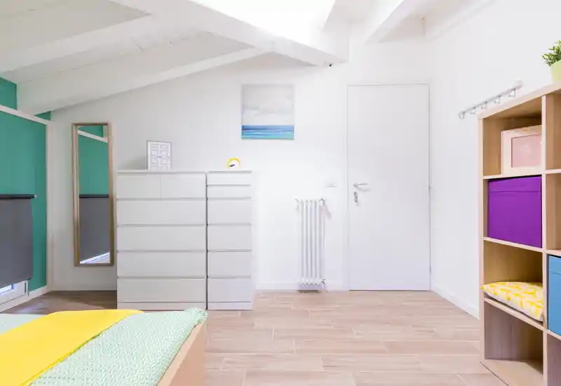 Bright, modern bedroom corner with a visible bed, white chest of drawers, mirror and organized shelving — airy attic-style ceiling and warm wooden floor.