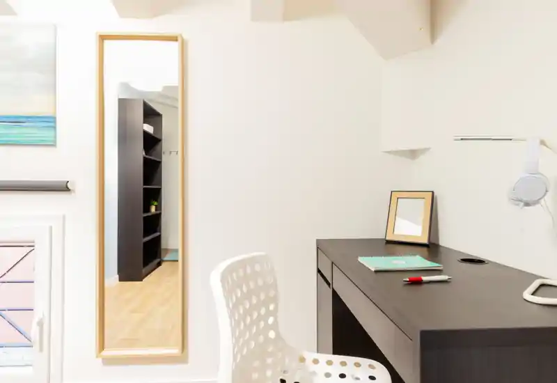 Bright, minimalist workspace corner within a living area: dark wood desk with a notebook and lamp, white perforated chair and a tall mirror that visually expands the space.