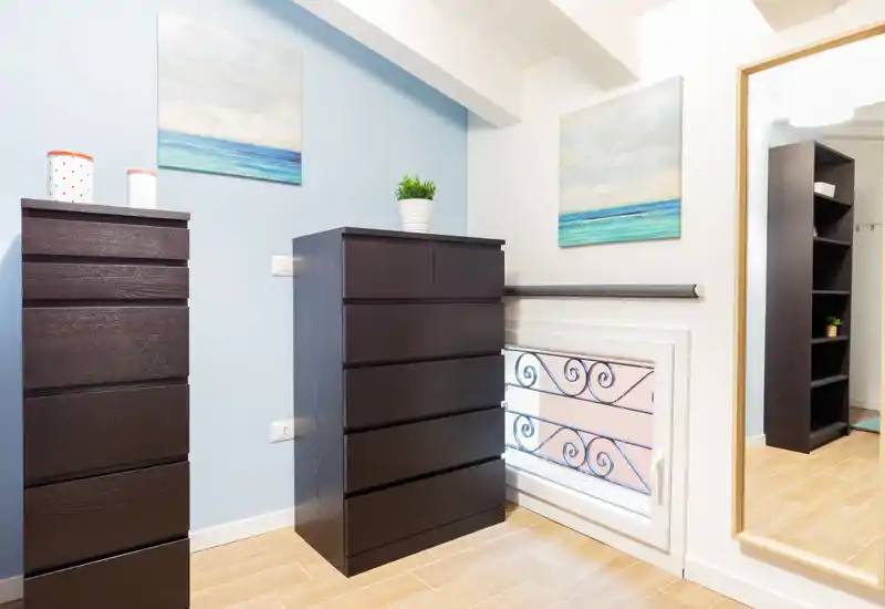 A small bedroom corner with two dark wood chests of drawers, decorative seascape paintings and a large mirror reflecting shelving. Warm wood flooring and a potted plant complete the bright, tidy space.