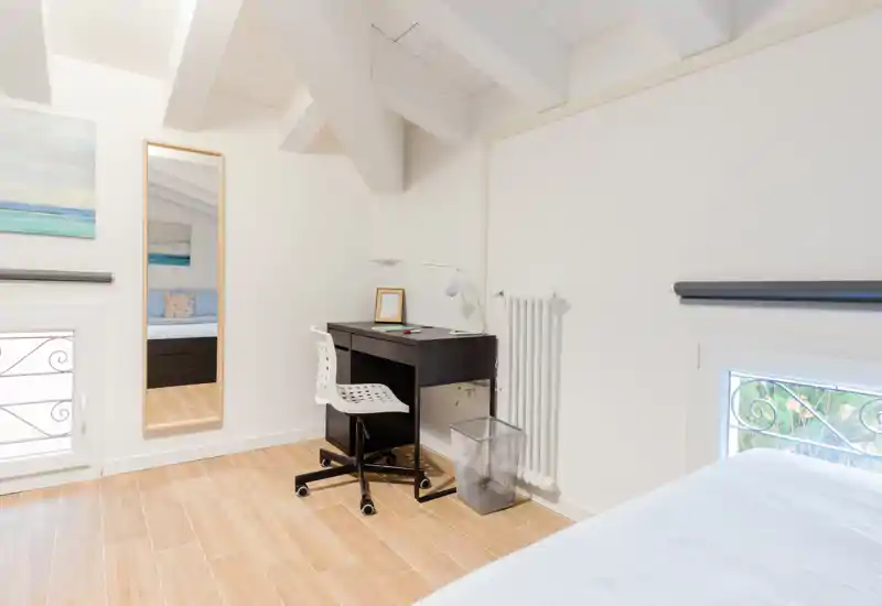 Bright, minimalist attic bedroom with a light wooden floor, a small black desk and chair, and a neatly made bed visible at the edge of the frame — ideal for single stays or short-term rentals.