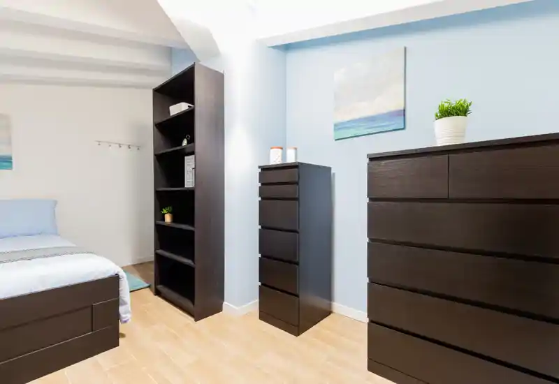 Dormitorio luminoso y ordenado con la cama visible, muebles oscuros a juego y cuadro de estilo costero, perfecto para un anuncio de alquiler.