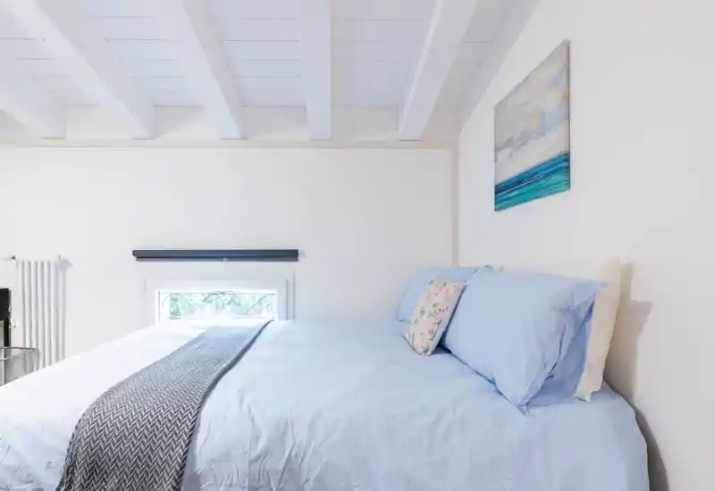 Dormitorio luminoso y aireado con una cama grande vestida en sábanas azul claro, cojines decorativos y un camino de cama texturizado — decoración minimalista y luz natural.