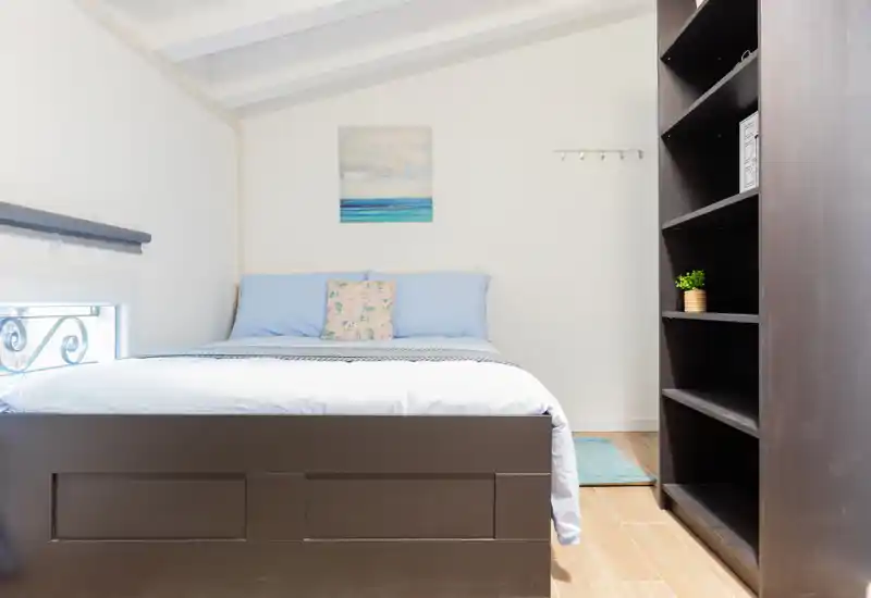 Dormitorio luminoso y bien decorado con cama doble, ropa de cama azul suave y cojín decorativo; una amplia estantería oscura al lado ofrece espacio de almacenaje.