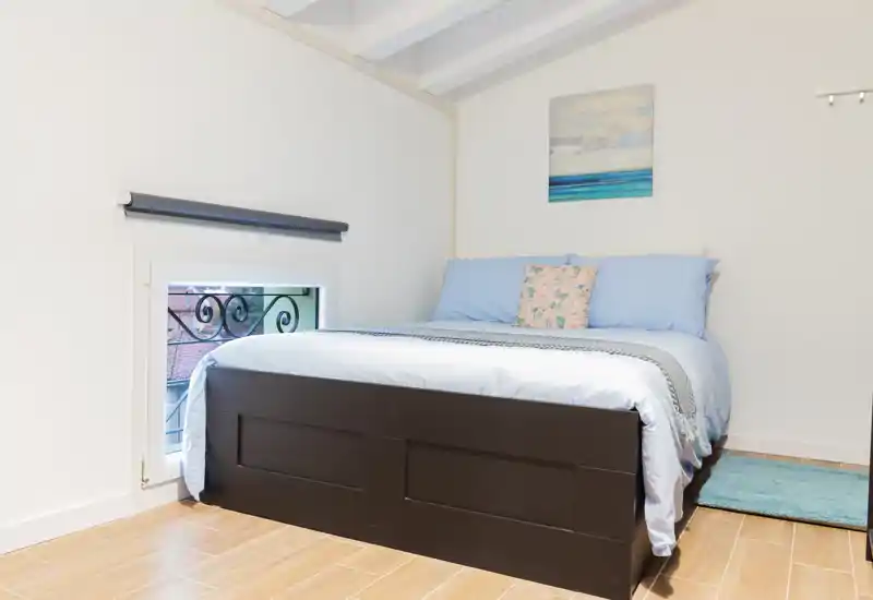 Dormitorio luminoso y ordenado con cama doble, ropa de cama azul clara, suelo de madera y un pequeño cuadro de inspiración costera.