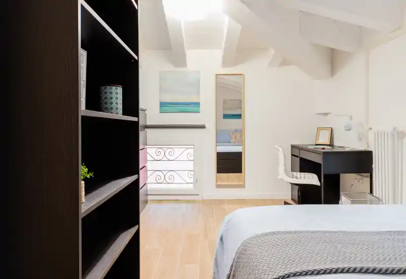 Camera da letto mansardata luminosa e moderna con letto visibile, pavimento in legno chiaro, scrivania e scaffalatura — stile minimal e curato, ottimo per un annuncio.