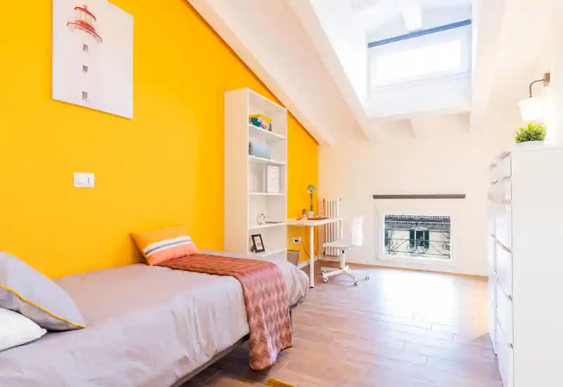 Dormitorio moderno y soleado con cama individual junto a una pared amarilla vibrante, escritorio junto a la ventana y luz natural intensa desde el tragaluz — ideal para estudiante o una persona.