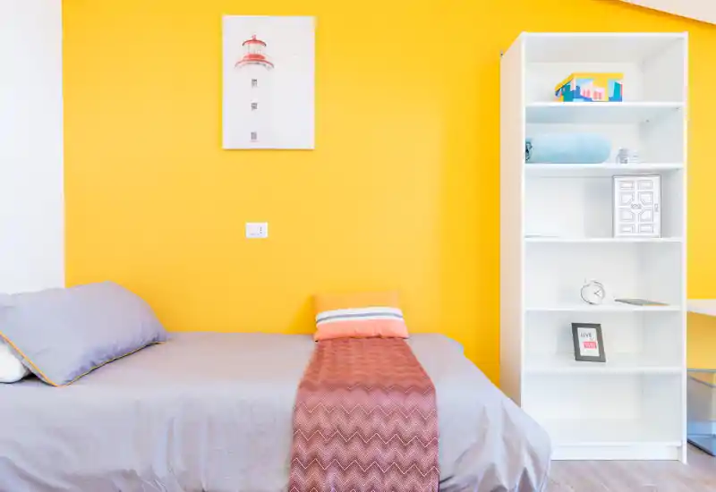 Dormitorio individual luminoso y bien presentado con edredón gris, plaid decorativo y estantería blanca frente a una pared amarilla vibrante — ideal para un alquiler moderno y acogedor.